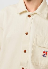 Camisa color crema con cuello, con botones de metal y un bolsillo. Incluye un logo en parche con una ilustración de un personaje.