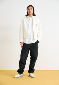 White cotton jacket with contrasting zipper, striped button-up shirt, black trousers, and light-colored sneakers, set against a plain background.