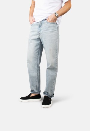 Person trägt hellblaue Jeans, weißes T-Shirt, schwarze Slipper und eine silberne Armbanduhr und steht mit den Händen in den Taschen.