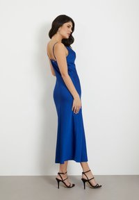 Woman in a fitted, sleeveless royal blue midi dress and black high-heeled sandals, standing against a white background.