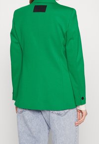 Blazer verde su misura con una texture lucida, dotato di un'etichetta nera sul retro e polsini con bottoni. Indossato con jeans azzurri chiari.