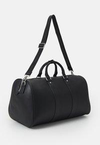 Black leather duffle bag with dual top handles, adjustable strap, silver hardware, and a zip closure. Features textured finish and logo strap.