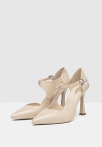 Escarpins en cuir beige à bout pointu, avec un design découpé sur les côtés, une bride de cheville avec un équipement en argent et des talons aiguilles fins.