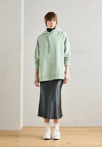Mintgroene oversized hoodie met een voorzak en een trekkoord, gecombineerd met een glanzende zwarte midi-jurk en witte sneakers.
