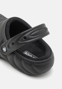 Zoccoli neri con superficie testurizzata, morbido rivestimento interno nero e una cinghia. Il design presenta forme arrotondate e un logo sulla cinghia.