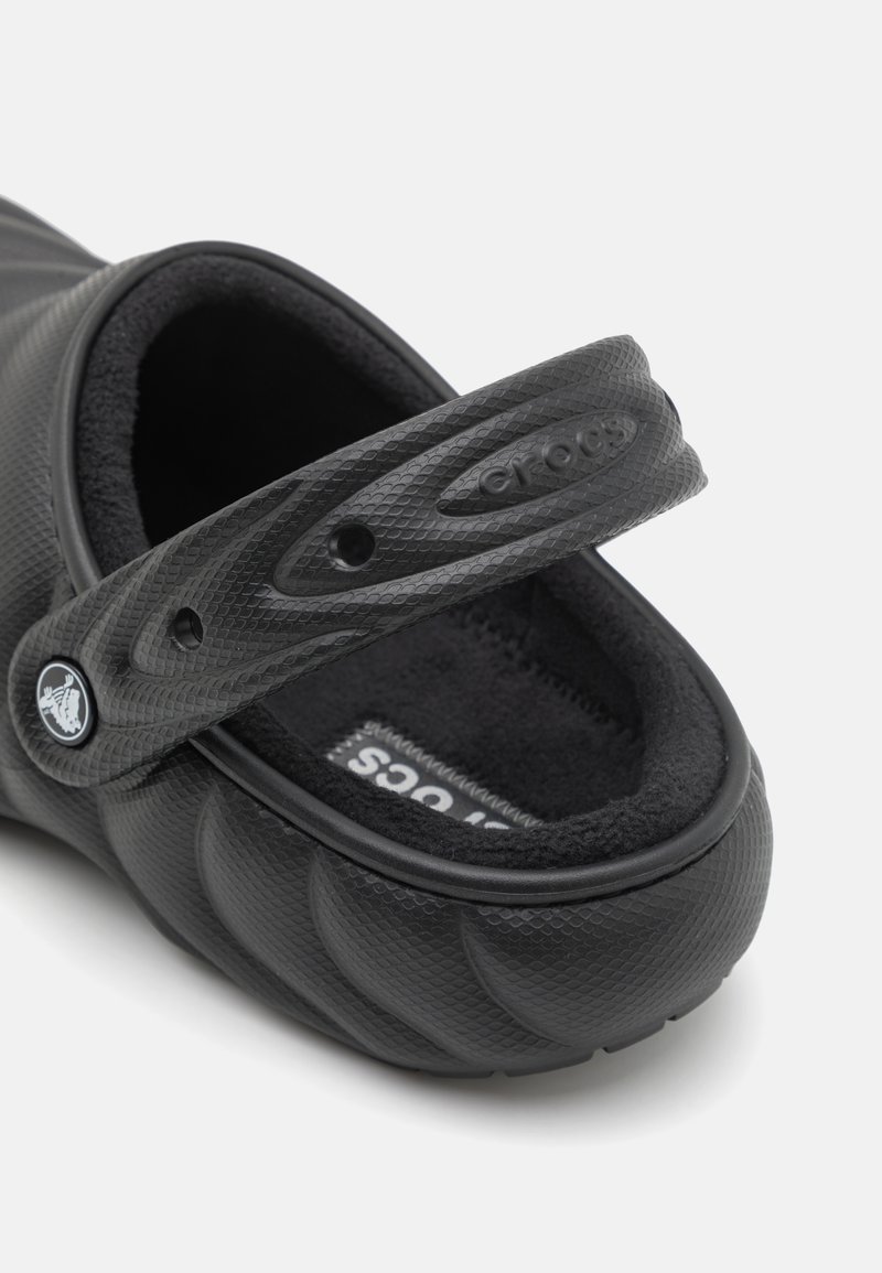 Zuecos negros con una superficie texturizada, un forro suave en negro y una correa. El diseño presenta formas redondeadas y un logotipo en la correa.