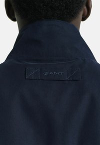Giacca blu navy con un colletto alto. Presenta un'etichetta con logo ricamato sul retro con accenti a punto croce. Tessuto dalla consistenza liscia.