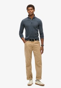 Mørkeblå langærmet polo-shirt, beige bomuldsbukser og hvide sneakers med lysegrå detaljer. Har en knappet krave og bælte.