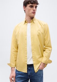 Άνδρας με κοντά καστανά μαλλιά, φοράει ανοιχτό κίτρινο πουκάμισο πάνω από λευκό μπλουζάκι και μπλε τζιν, με το ένα χέρι στην τσέπη, κοιτάζοντας δεξιά.