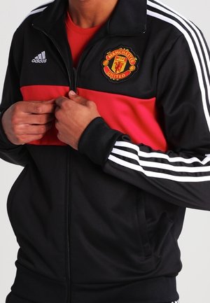Personne portant une veste Adidas Manchester United noire et rouge avec trois bandes blanches sur les manches, en train de fermer la fermeture éclair.
