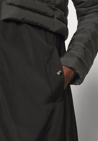 Chaqueta acolchada negra con textura acolchada y un bolsillo lateral con un botón a presión metálico redondo. La manga tiene un acabado suave.