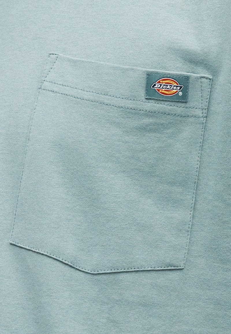 Tasca in tessuto verde chiaro con una toppa rettangolare cucita che presenta il logo del marchio Dickies in rosso, arancione, nero e bianco.