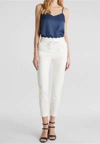 Femme portant un haut sans manches bleu marine, un pantalon blanc taille haute avec une taille festonnée et des talons noirs à brides, debout sur fond gris.