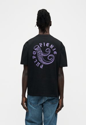 Άνδρας με κοτσιδάκια, φοράει μαύρο μπλουζάκι με μοβ πλοκάμι χταποδιού και το κείμενο "PULPO PICNIC" στην πλάτη, συνδυασμένο με μπλε τζιν.