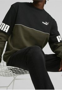 Sudadera negra y verde oliva de manga larga, que presenta un logo blanco y detalles de ribete en el pecho, sentada sobre una silla blanca.