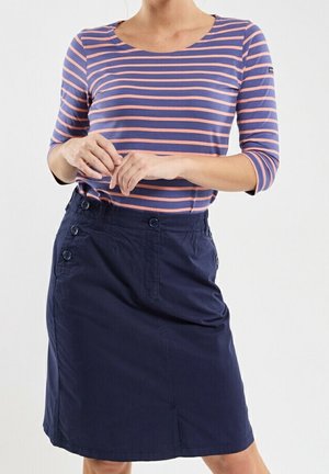 Kvinde iført en blå nederdel med knap detaljer og en lilla langærmet top med lyserøde vandrette striber, hænderne foldet foran.