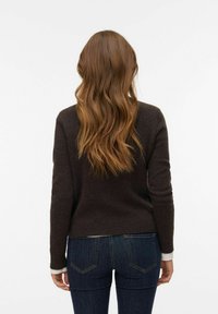 Brauner, gestrickter Pullover mit langen Ärmeln, abgerundetem Saum und kontrastierenden hellen Bündchen, kombiniert mit engen dunklen Jeans. Glatte Textur, lässiges Design.