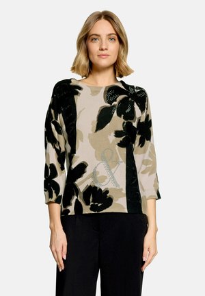 Femme aux cheveux châtain clair portant un pull beige avec de grands motifs floraux noirs et un pantalon noir, debout devant un fond uni.
