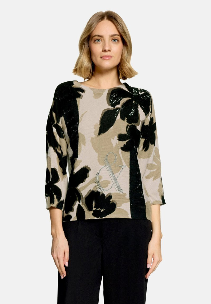 Femme aux cheveux châtain clair portant un pull beige avec de grands motifs floraux noirs et un pantalon noir, debout devant un fond uni.