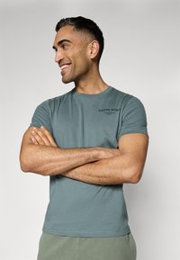 T-shirt in cotone color teal con colletto rotondo, maniche corte e logo stampato in nero sul petto. Texture liscia, design casual.