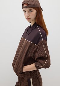 Bruine geruite blouse met een ontspannen fit, voorzien van een donkerbruine schouderpas, contrasterende lichte piping en elastische manchetten. Draagt met een bruine pet.