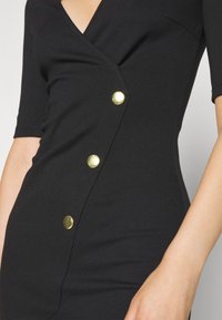 Robe noire ajustée avec un décolleté en V profond, des manches courtes et une patte de boutonnage diagonale ornée de quatre boutons dorés sur le côté.