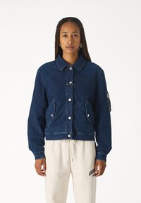 Giubbotto in denim di colore blu scuro con colletto, bottoni a pressione frontali, due tasche laterali e una tasca aggiuntiva sulla manica sinistra.