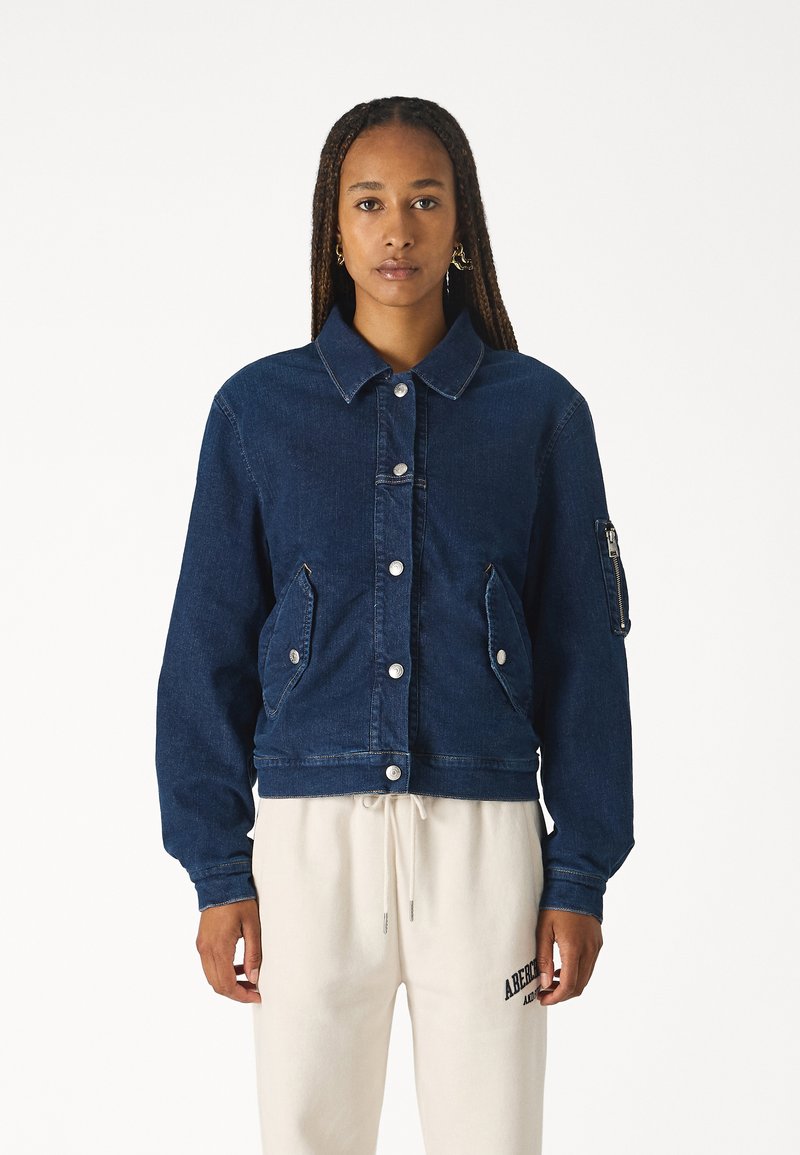 Giubbotto in denim di colore blu scuro con colletto, bottoni a pressione frontali, due tasche laterali e una tasca aggiuntiva sulla manica sinistra.