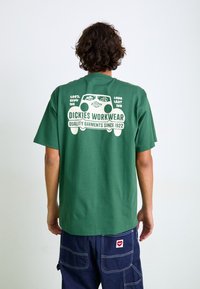 Grünes Baumwoll-T-Shirt mit einem weißen grafischen Design auf der Rückseite, das Text und Bilder enthält. Kurze Ärmel, lockere Passform, lässiger Stil.