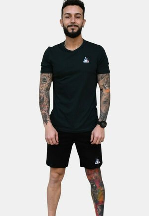 Camiseta negra con un pequeño logo, combinada con pantalones cortos negros. Presenta mangas cortas, un diseño ajustado y tatuajes visibles en los brazos y las piernas.