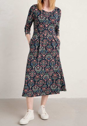 Donna che indossa un abito midi blu navy con motivi floreali rosa, rossi e bianchi, maniche a tre quarti, tasche e sneakers bianche alte.