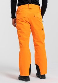 Heldere oranje skibroek met een gladde textuur, verstelbare banden, meerdere zakken en zwarte accenten bij de enkel voor extra duurzaamheid.