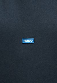 Μπλε ορθογώνιο ένθετο με λευκό κείμενο "HUGO" ραμμένο σε σκουρόχρωμο γκρι ύφασμα.