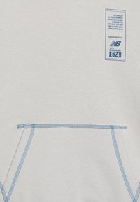 Jasnoszara bluza z niebieskimi przeszyciami na kieszeni i dolnym obszyciu. Posiada niebieską metkę z danymi produktu w lewym górnym rogu.