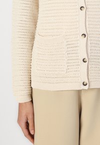 Beige neulejacke, jossa on avoin kudontamalli, etupainikkeet ja yksi tasku. Päällä vaaleanbeige housut. Pehmeä rakenne, rento istuvuus.