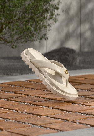 Sandale tip șlapi bej, cu talpă groasă și pernată și fundul zimțat, așezată în unghi pe o suprafață de lemn cu plăci, în aer liber.