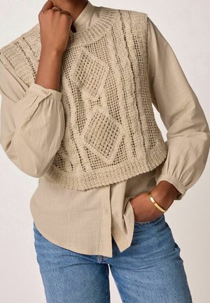 Persoon draagt een beige gehaakte mouwloze trui over een beige blouse met lange mouwen en knopen, gecombineerd met blauwe jeans en een gouden armband.