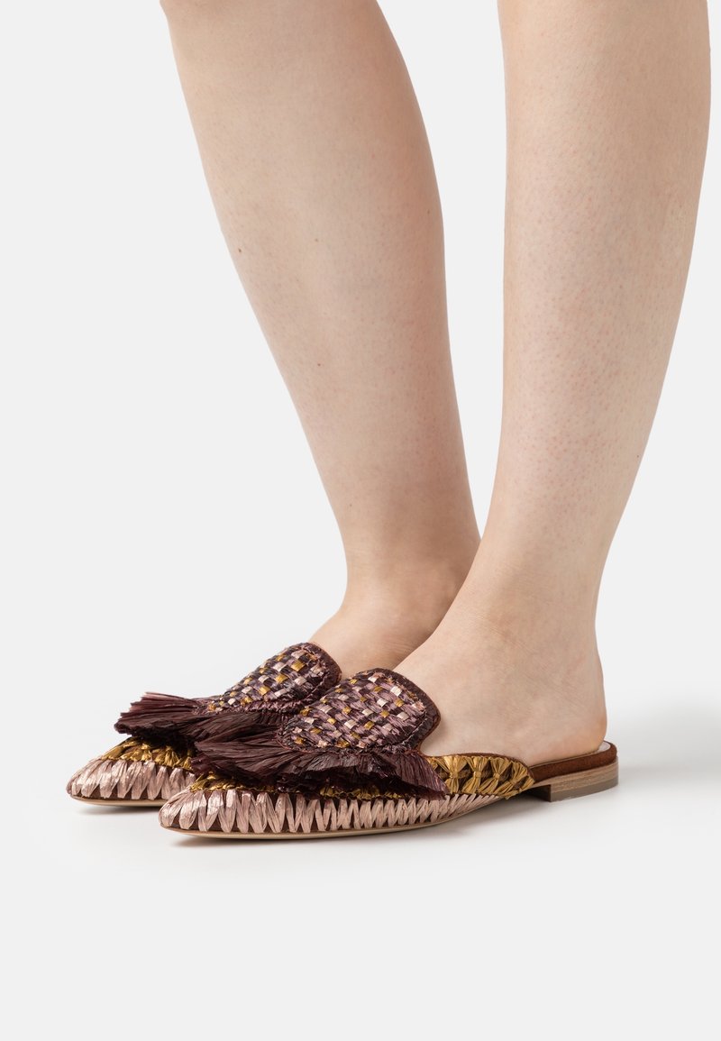 Pieds chaussés de mocassins plats tissés avec des bouts pointus et de grands pompons marron foncé sur la partie avant supérieure.