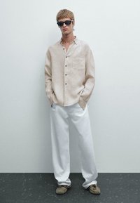 Beige Leinen-Hemd mit Knopfverschluss und Brusttasche, kombiniert mit lockeren weißen Hosen und braunen Slippern, getragen von einem Model.