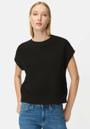 Kvinde med glat blondt hår iført en sort ribbet kortærmet sweater og blå jeans, stående foran en ensfarvet lys baggrund.