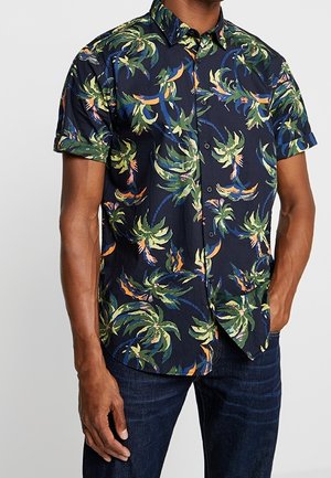 Mand iført en kortærmet skjorte med knapper, dekoreret med et farverigt tropisk palmemønster, og mørkeblå jeans.