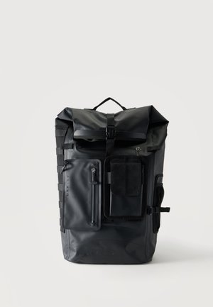 Zaino nero con chiusura roll-top, multiple tasche frontali con zip, chiusura a fibbia e maniglia laterale su uno sfondo chiaro uniforme.