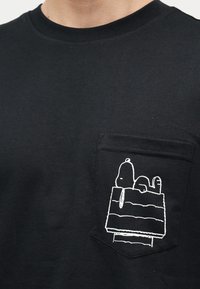 Μαύρο T-shirt με τσεπάκι στο στήθος που απεικονίζει με λευκή γραμμή ένα σκύλο να ξαπλώνει πάνω σε ένα σπιτάκι για σκύλους.