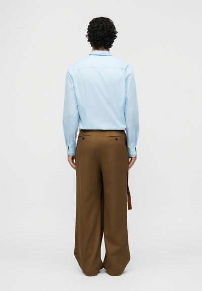 Homme aux cheveux bouclés et foncés portant une chemise habillée bleu clair rentrée dans un pantalon large marron, vu de dos sur fond blanc.