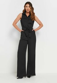 Svart ärmlös jumpsuit med knäppning, insvängd midja med knytband och vida byxor, med en mjuk yta och skräddarsydd design.