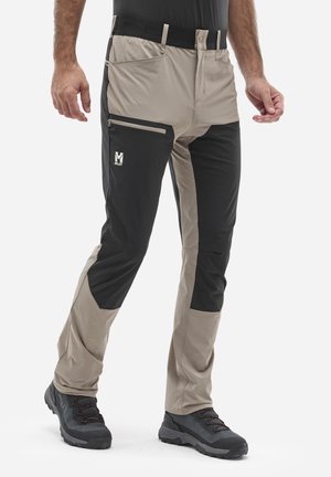 Man wearing beige and black hiking pants with zippered thigh pocket and black hiking shoes on white background.