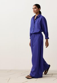 Frau geht in locker sitzendem, lila Langarmshirt und passenden, weit geschnittenen Hosen mit schwarzen Flip-Flops auf hellem Fliesenboden.