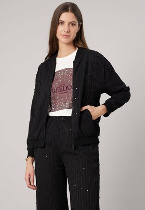 Vrouw die een zwarte, gestructureerde jas en broek draagt met een wit t-shirt met een paarse afbeelding met de tekst "Freedom" op een neutrale achtergrond.