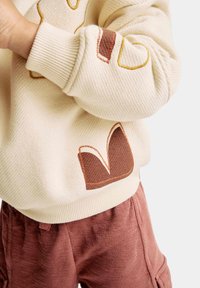 Stickad krämfärgad sweatshirt med broderade bruna och gula detaljer, med en texturerad mönster och ribbade manschetter; matchad med rostfärgade byxor.