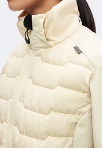 Femme portant une veste matelassée crème avec un col haut et un cordon noir réglable près du cou, montrant le haut du torse et l’épaule.